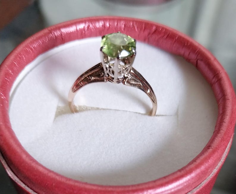 May include: A gold-toned ring with a light green gemstone, set in a detailed filigree design. The ring is presented in a red ring box with a white interior, showcasing the jewelry's elegance. The gemstone is round and faceted.
