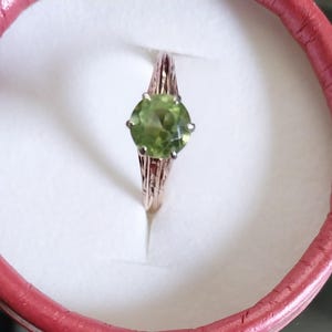 May include: A gold-colored ring with a large, round, light green gemstone. The ring is set in a decorative band and is displayed inside a pink ring box.