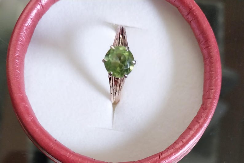 May include: A gold-colored ring with a large, round, light green gemstone. The ring is set in a decorative band and is displayed inside a pink ring box.