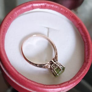 May include: A gold-toned ring with a light green gemstone set in a decorative cage setting. The ring is displayed inside a red, round box with a white interior, showcasing the jewelry's intricate design and color contrast.