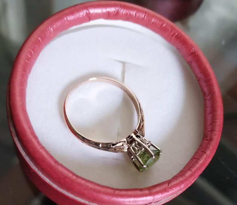 May include: A gold-toned ring with a light green gemstone set in a decorative cage setting. The ring is displayed inside a red, round box with a white interior, showcasing the jewelry's intricate design and color contrast.