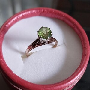May include: A gold ring with a large, round, light green gemstone. The ring is set in a detailed, filigree design and is presented in a red ring box with a white interior.