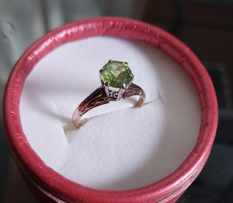 May include: A gold ring with a large, round, light green gemstone. The ring is set in a detailed, filigree design and is presented in a red ring box with a white interior.