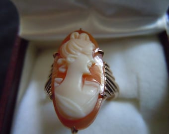 May include: Oval cameo ring featuring a profile of a woman carved in cream-colored relief against an orange background. The ring has a gold-colored setting with decorative elements on the sides. The ring is displayed inside a jewelry box.