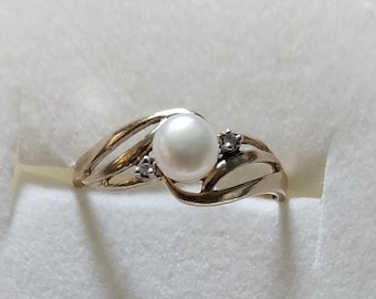 May include: A gold-toned ring featuring a large, round, white pearl as the centerpiece. The ring has a flowing, open design with two smaller, clear stones on either side of the pearl. The ring is set against a white background.