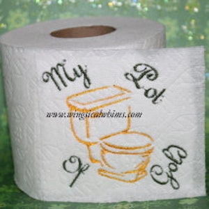 Pode incluir: Um rolo de papel higiênico branco com um desenho bordado em dourado que diz "My Pot of Gold" e inclui um vaso sanitário e uma escova de vaso sanitário.