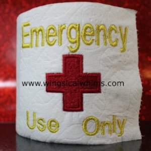 May include: White toilet paper roll with the words "Emergency Use Only" embroidered in yellow thread. A red embroidered cross is centred on the roll. The website www.wingslicalwhims.com is printed below the cross.