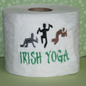 Op de afbeelding: Een witte toiletrol met een groene, bruine en zwarte illustratie van drie stokpoppetjes in verschillende yogaposes. De tekst "IRISH YOGA" is in het groen onder de poppetjes gedrukt.