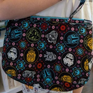 May include: Black tote bag with a colorful pattern featuring Star Wars characters and Day of the Dead sugar skulls. The bag has a blue interior and a matching strap. The bag is held by a person.