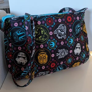 May include: Black tote bag with a colorful pattern featuring Star Wars characters and sugar skull designs. The bag has a blue zipper and straps, and is made of a durable fabric. The bag is designed for carrying items.