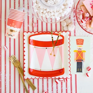 Puede incluir: Suministros para fiestas festivas sobre un fondo a rayas rojas y blancas. Incluye un plato en forma de tambor con baquetas doradas, una tarjeta de cascanueces, tenedores dorados y vasos con diseño de bigote. Caramelos de menta y dulces esparcidos.