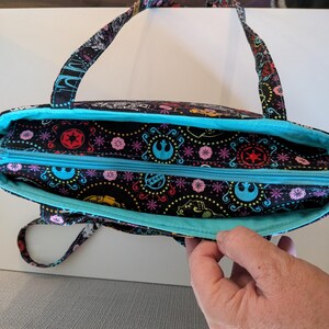 Puede incluir: Bolso tote negro con interior y cremallera turquesa. El bolso presenta un colorido estampado de s&iacute;mbolos y dise&ntilde;os de Star Wars. Las asas y la cremallera son turquesas. El bolso est&aacute; abierto, revelando el interior.