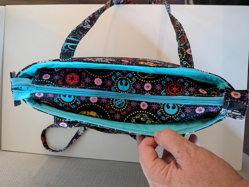 Puede incluir: Bolso tote negro con interior y cremallera turquesa. El bolso presenta un colorido estampado de s&iacute;mbolos y dise&ntilde;os de Star Wars. Las asas y la cremallera son turquesas. El bolso est&aacute; abierto, revelando el interior.