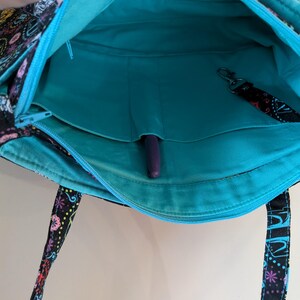 Puede incluir: Vista abierta de un bolso tote negro con un colorido estampado floral. El interior es turquesa con un bolsillo con cremallera y un portal&aacute;pices. Se adjunta un cord&oacute;n negro con un clip. Las asas de la bolsa tienen un estampado.