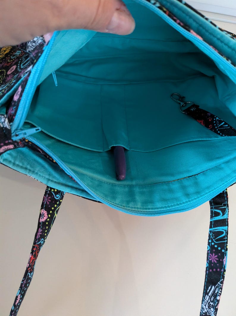 Puede incluir: Vista abierta de un bolso tote negro con un colorido estampado floral. El interior es turquesa con un bolsillo con cremallera y un portal&aacute;pices. Se adjunta un cord&oacute;n negro con un clip. Las asas de la bolsa tienen un estampado.