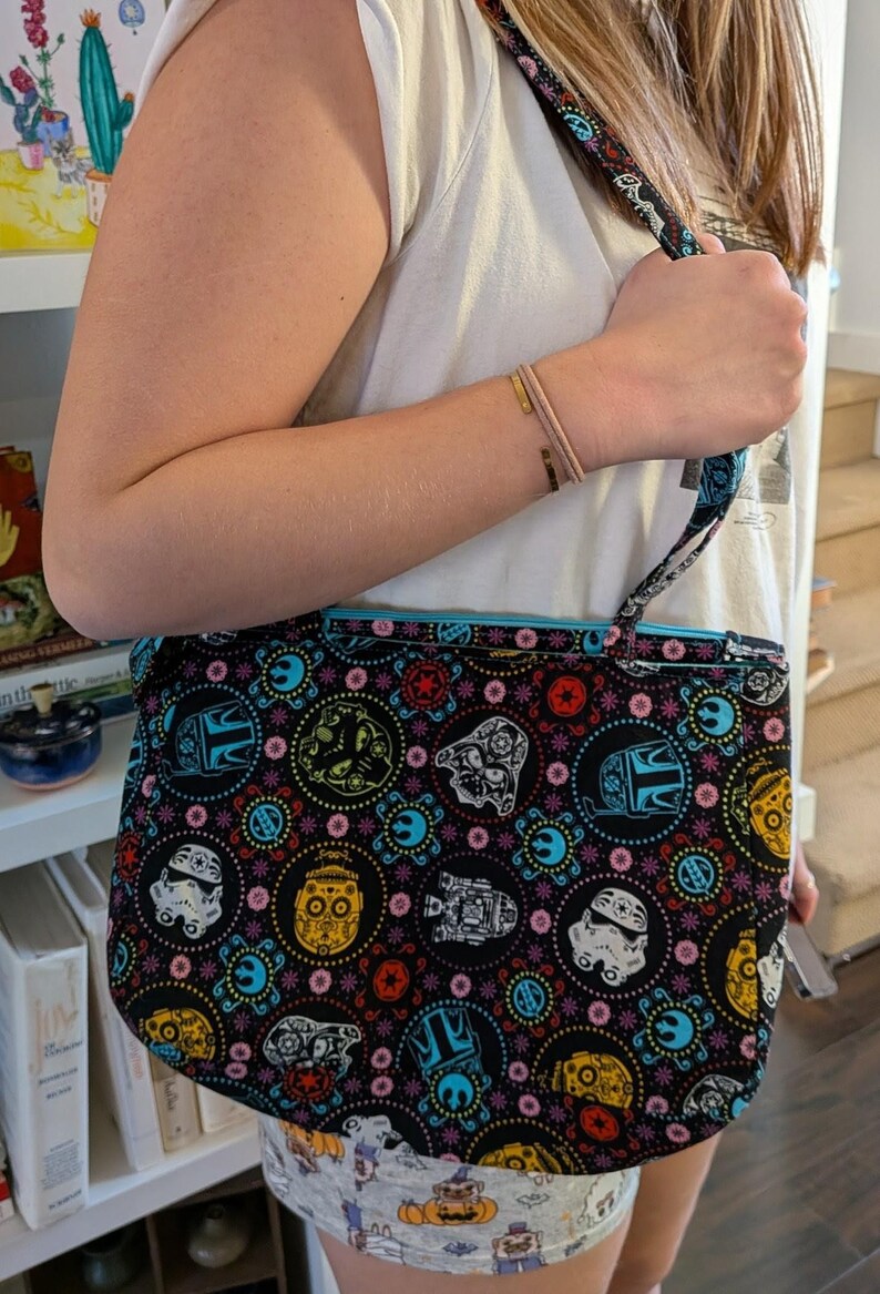 May include: Black tote bag with a colorful pattern featuring Star Wars characters and Day of the Dead sugar skulls. The bag has a blue interior and a matching strap. The bag is held by a person.