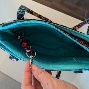 Puede incluir: Bolso tote abierto con fondo negro y estampado floral colorido. El interior es turquesa y el bolso tiene una cremallera turquesa. Una peque&ntilde;a correa con un cierre plateado est&aacute; sujeta en el interior. El bolso tiene dos asas.
