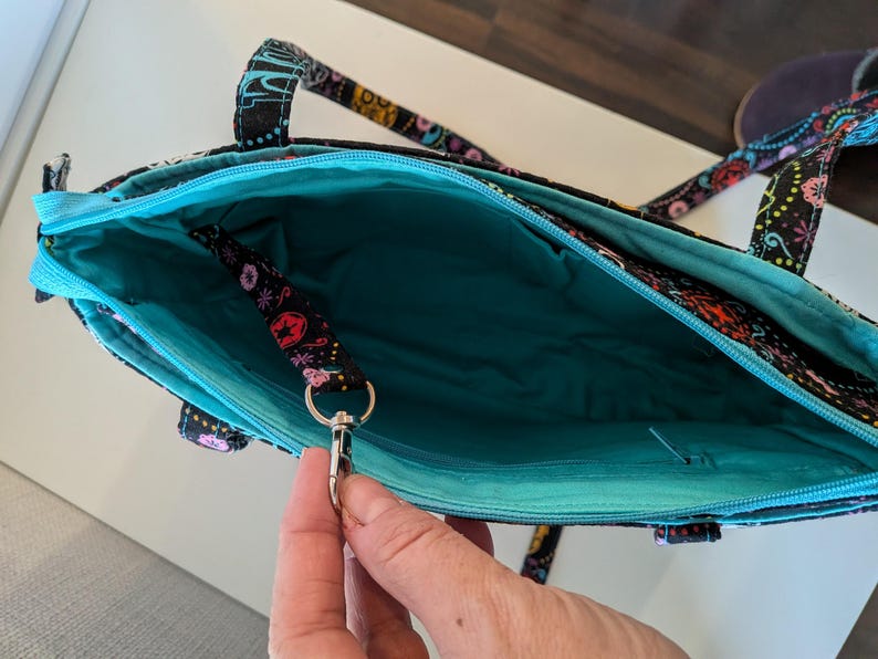 Puede incluir: Bolso tote abierto con fondo negro y estampado floral colorido. El interior es turquesa y el bolso tiene una cremallera turquesa. Una peque&ntilde;a correa con un cierre plateado est&aacute; sujeta en el interior. El bolso tiene dos asas.