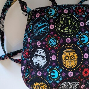 Puede incluir: Bolso tote negro con un patr&oacute;n colorido con personajes de Star Wars y dise&ntilde;os de calaveras de az&uacute;car del D&iacute;a de Muertos. El bolso tiene un interior azul y correas a juego. El dise&ntilde;o incluye un casco de Stormtrooper, un casco mandaloriano y otros s&iacute;mbolos ic&oacute;nicos.
