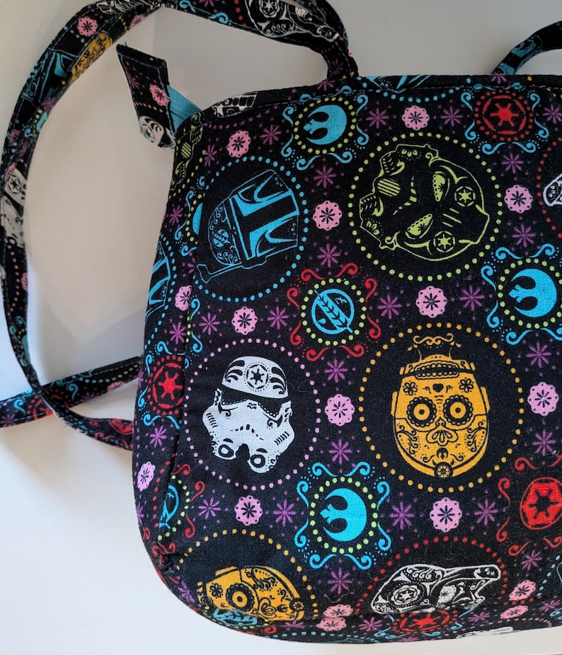Puede incluir: Bolso tote negro con un patr&oacute;n colorido con personajes de Star Wars y dise&ntilde;os de calaveras de az&uacute;car del D&iacute;a de Muertos. El bolso tiene un interior azul y correas a juego. El dise&ntilde;o incluye un casco de Stormtrooper, un casco mandaloriano y otros s&iacute;mbolos ic&oacute;nicos.