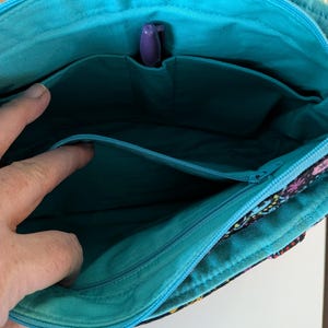 Puede incluir: Vista interior de un bolso tote con estampado floral y un interior turquesa. El bolso tiene m&uacute;ltiples bolsillos y un portal&aacute;pices morado. La cremallera es turquesa. El exterior del bolso presenta un fondo negro con dise&ntilde;os florales coloridos.