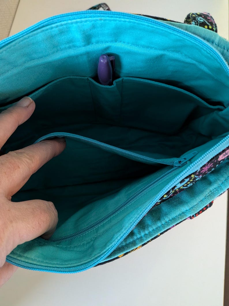 Puede incluir: Vista interior de un bolso tote con estampado floral y un interior turquesa. El bolso tiene m&uacute;ltiples bolsillos y un portal&aacute;pices morado. La cremallera es turquesa. El exterior del bolso presenta un fondo negro con dise&ntilde;os florales coloridos.