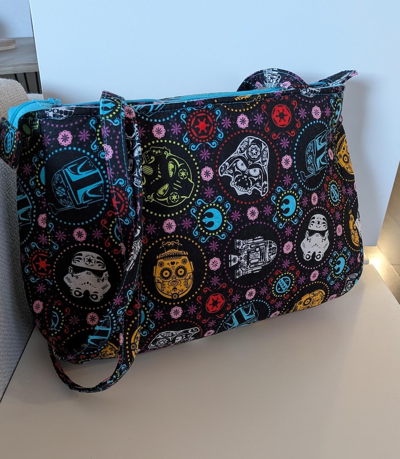 May include: Black tote bag with a colorful pattern featuring Star Wars characters and sugar skull designs. The bag has a blue zipper and straps, and is made of a durable fabric. The bag is designed for carrying items.