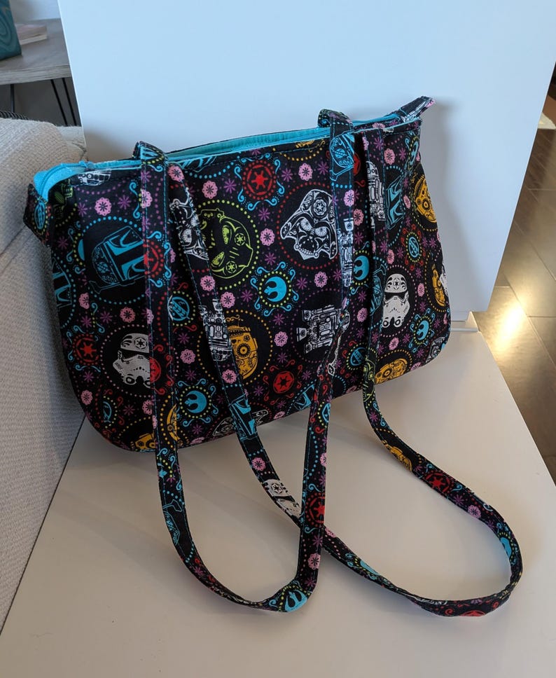 Puede incluir: Bolso tote negro con un colorido estampado de Star Wars. El bolso presenta im&aacute;genes de Stormtroopers, Darth Vader y otros personajes ic&oacute;nicos. El interior est&aacute; forrado con una tela azul brillante y el bolso tiene asas a juego.