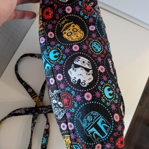 Puede incluir: Bolso de tela negra con un patr&oacute;n colorido con personajes de Star Wars y calaveras de az&uacute;car del D&iacute;a de Muertos. El dise&ntilde;o incluye Stormtroopers, Boba Fett y otros s&iacute;mbolos ic&oacute;nicos, con una correa a juego.