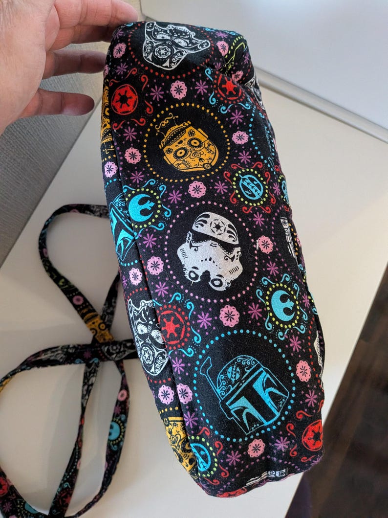 Puede incluir: Bolso de tela negra con un patr&oacute;n colorido con personajes de Star Wars y calaveras de az&uacute;car del D&iacute;a de Muertos. El dise&ntilde;o incluye Stormtroopers, Boba Fett y otros s&iacute;mbolos ic&oacute;nicos, con una correa a juego.