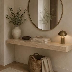 May include: A floating beige stone console table holds a white vase with green branches, a dark bowl, two stacked books, a small dish, and a brass mushroom lamp. A round mirror hangs above, and a woven basket with a beige cloth sits below.