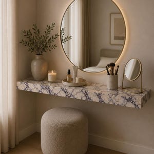 May include: A floating vanity shelf with a purple and white marble pattern, paired with a round backlit wall mirror, a beige textured ottoman, a ceramic vase with greenery, a candle, a small tray, a brush holder, and a gold tabletop mirror.