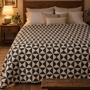 May include: A bed quilt featuring a repeating geometric pattern of black and white triangles arranged in squares. The quilt covers the bed, which is styled with several white and beige pillows, including a long rectangular lumbar pillow.