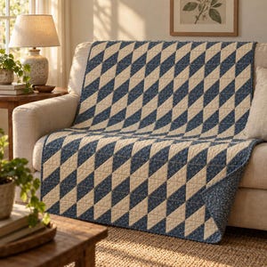Op de afbeelding: Een rechthoekige quilt met een herhalend geometrisch patroon van afwisselende crèmekleurige driehoeken en donkerblauwe driehoeken met een kleine witte bloemenprint. De quilt ligt over een bank en toont gestikte naden.