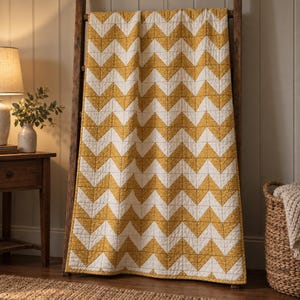 Peut inclure: Une courtepointe rectangulaire présentant un motif à chevrons répétitif en jaune moutarde et blanc cassé. Les sections jaunes affichent un subtil imprimé floral, tandis que les sections blanches sont unies. La courtepointe est drapée sur une échelle en bois.
