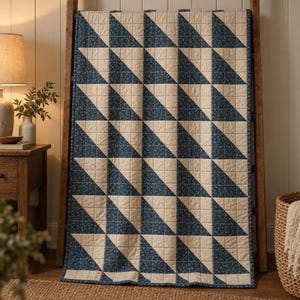 May include: A rectangular quilt featuring a repeating half-square triangle pattern. The design alternates between solid cream-colored fabric and a dark blue fabric with a small, intricate white floral print throughout the textile.