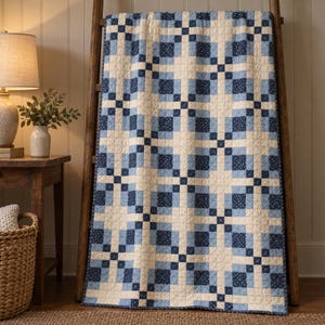 May include: A rectangular quilt featuring a repeating grid pattern of cream, light blue, and dark navy blue squares. Some dark blue squares contain a small white floral print. The quilt is draped over a wooden ladder frame.