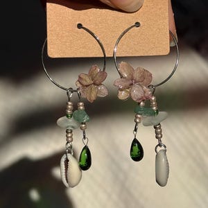 Könnte beinhalten: Ein Paar Creolen mit befestigten getrockneten rosa Hortensienblüten, kleinen Metallperlen, durchscheinendem grünem Meerglas, dunkelgrünen tropfenförmigen Glasanhängern und weißen Kaurimuscheln, die an dünnen silberfarbenen Metallreifen hängen.