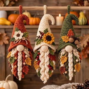 May include: Three hanging crochet gnomes in autumn colors. The left gnome is burnt orange, the center is cream, and the right is forest green. Each features a pointed hat, a round nose, a braided beard, and decorative felt leaves and berries.
