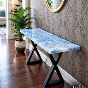 May include: A rectangular console table featuring a polished blue and white stone top with a marbled appearance. The table is supported by two black metal legs arranged in an X-shaped design. The logo LuxeWoodNStone is visible in the corner.