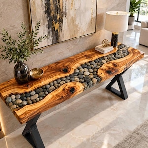 Puede incluir: Una mesa consola rectangular con un tablero de madera de borde natural y una incrustación central de piedras de río grises y negras lisas encapsuladas en resina transparente, sostenida por dos patas de metal negro en forma de X.