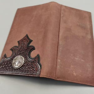 May include: A brown leather checkbook cover featuring a dark brown, intricately tooled leather corner overlay with a basketweave pattern and a central silver-toned circular concho ornament embossed with a five-pointed star design.
