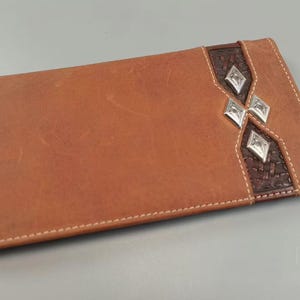 May include: A rectangular tan leather checkbook cover featuring white contrast stitching. The right side includes a dark brown textured leather inlay decorated with three silver-toned diamond-shaped metal studs arranged vertically.
