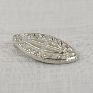 May include: A silver oval-shaped pendant with a raised design of a figure with a halo. The pendant has a decorative border.