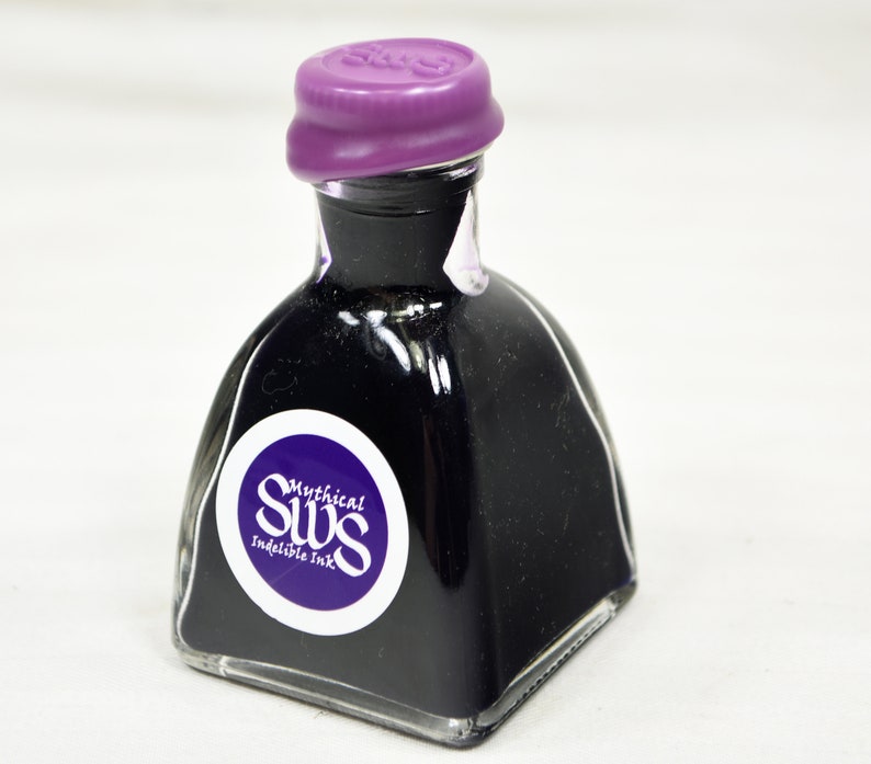 K&ouml;nnte beinhalten: Kleine, klare Glas-Tintenflasche gef&uuml;llt mit dunkelvioletter Tinte. Die Flasche hat einen lila Verschluss und ein wei&szlig;es und lila Etikett mit der Aufschrift "Mythical SWS Indelible Ink". Die Flasche hat eine einzigartige, quadratische Form.