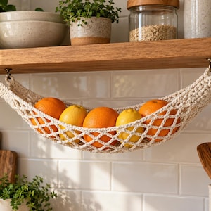 Puede incluir: Una hamaca para frutas de macramé color crema suspendida de un estante de madera mediante cuatro ganchos metálicos. La red tejida contiene tres naranjas brillantes y dos limones amarillos, con un patrón de rombos en toda la estructura.