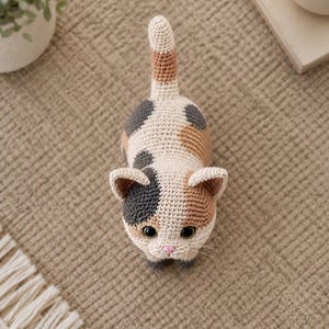 Peut inclure: Une petite figurine de chat calico crochetée avec une base crème et des taches gris foncé et marron clair. Le chat a de grands yeux noirs ronds, un petit nez rose brodé et une longue queue dressée avec des rayures marron et crème.