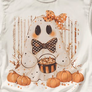Puede incluir: Una camiseta blanca con el gráfico de un fantasma que lleva una pajarita de lunares blancos y negros y un lazo para el pelo de lunares naranjas y blancos. El fantasma sostiene un cubo de dulces a rayas, rodeado de calabazas naranjas.