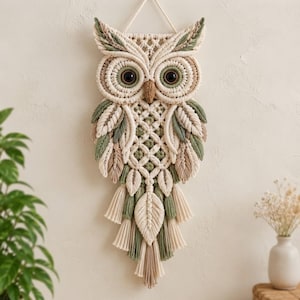 May include: A macrame wall hanging shaped like an owl, crafted from cream, sage green, and tan cotton cord. The owl features large dark brown eyes, a textured beak, and intricate knotting patterns forming wings and feathers with tassels.