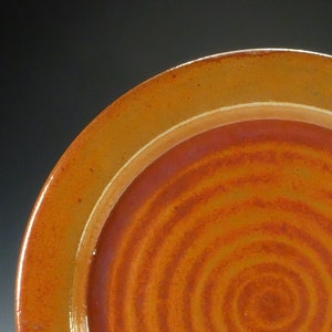 May include: A round ceramic plate with a swirling, concentric pattern in shades of orange and brown. The plate has a raised rim.
